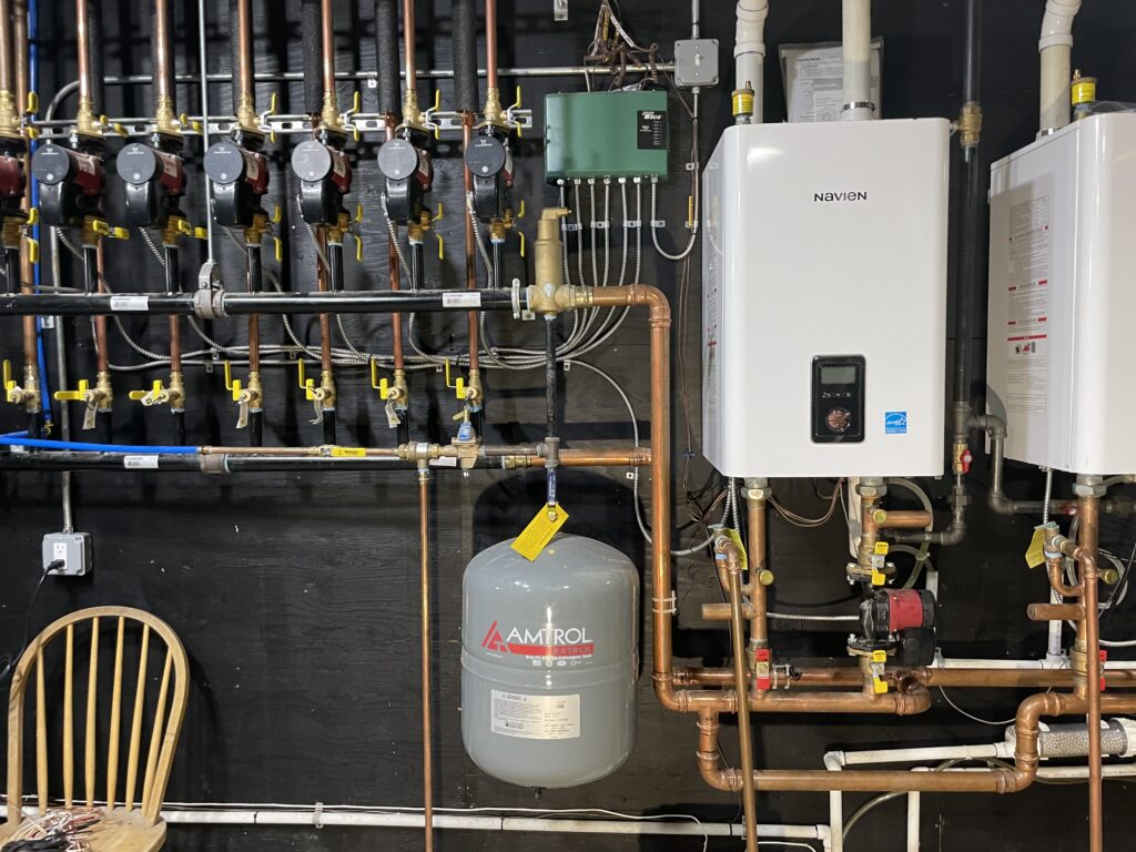 Wall mounted Navien boilers with a bank of circulator pumps, zone controls and an expansion tank in a mechanical room, installed by Action Construction